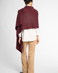 A woman shown from the back wearing a deep burgundy draped wrap or shawl over a white blouse, tan pants, and black shoes, against a light gray studio background.