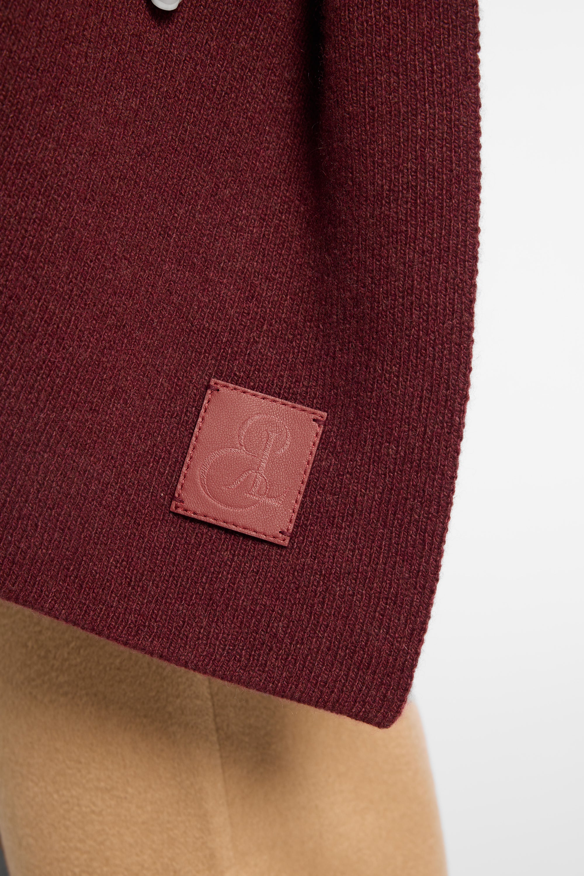 Close-up of a knit wrap in deep burgundy with a small square embossed leather patch, shown against a light neutral background.