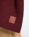 Close-up of a knit wrap in deep burgundy with a small square embossed leather patch, shown against a light neutral background.