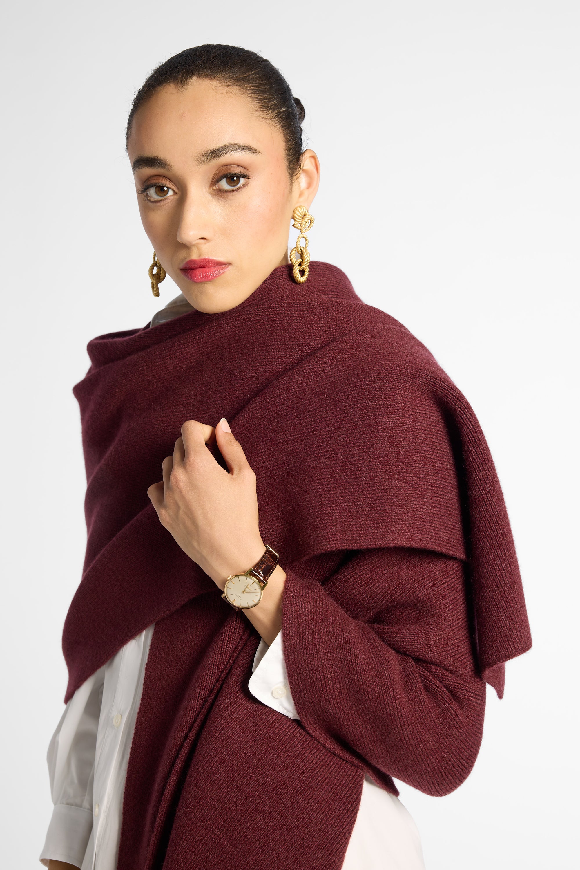 A woman wearing a chunky knit burgundy shawl draped over her shoulders, styled over a white top, against a plain light gray studio background.