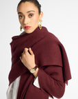 A woman wearing a chunky knit burgundy shawl draped over her shoulders, styled over a white top, against a plain light gray studio background.