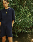 A woman wearing a dark navy blue knit sweater and matching shorts stands outdoors near a stream with dense green foliage in the background.