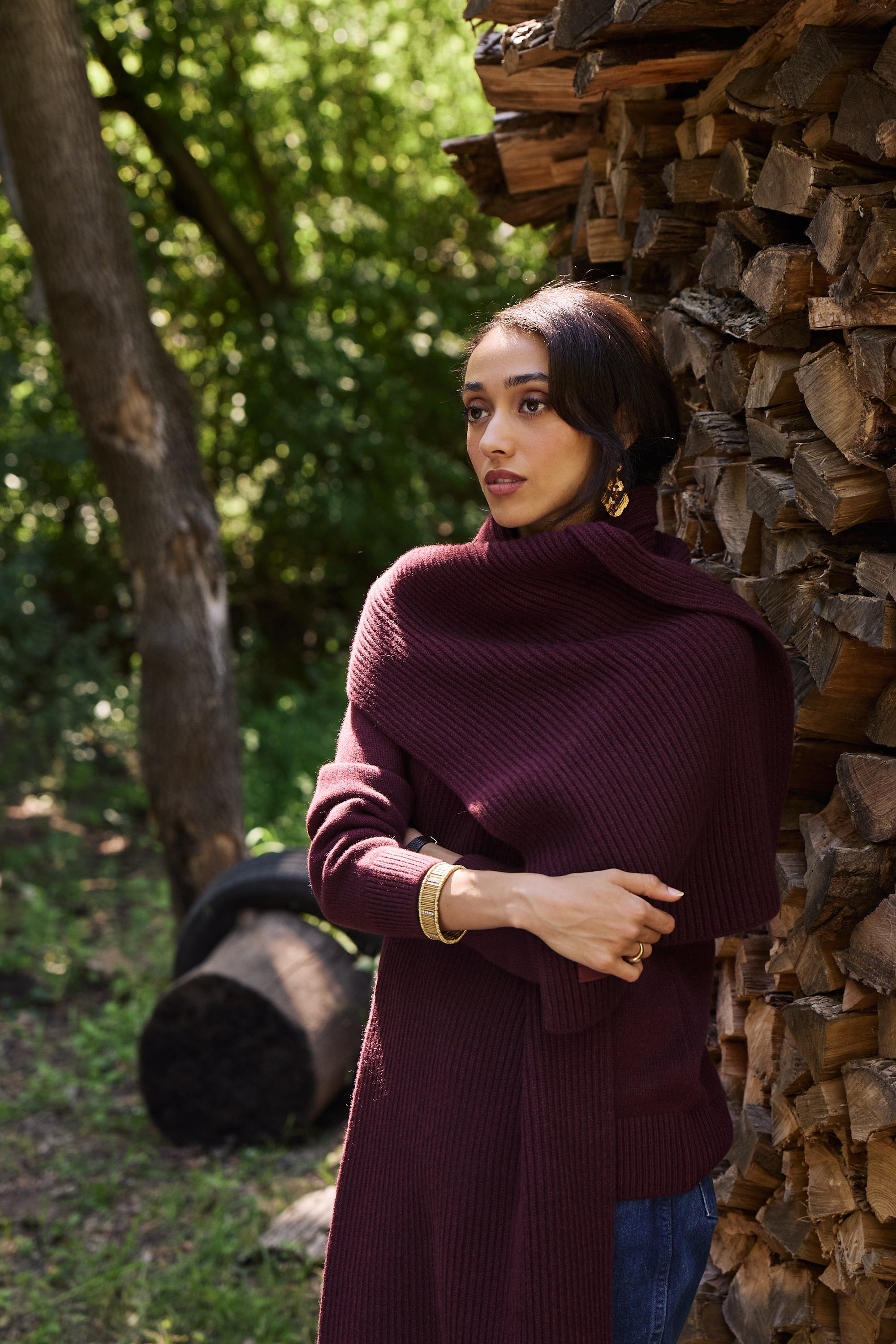 A woman wearing a ribbed knit turtleneck sweater in deep burgundy, standing outdoors next to a stacked woodpile.