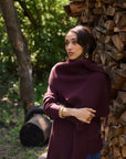 A woman wearing a ribbed knit turtleneck sweater in deep burgundy, standing outdoors next to a stacked woodpile.