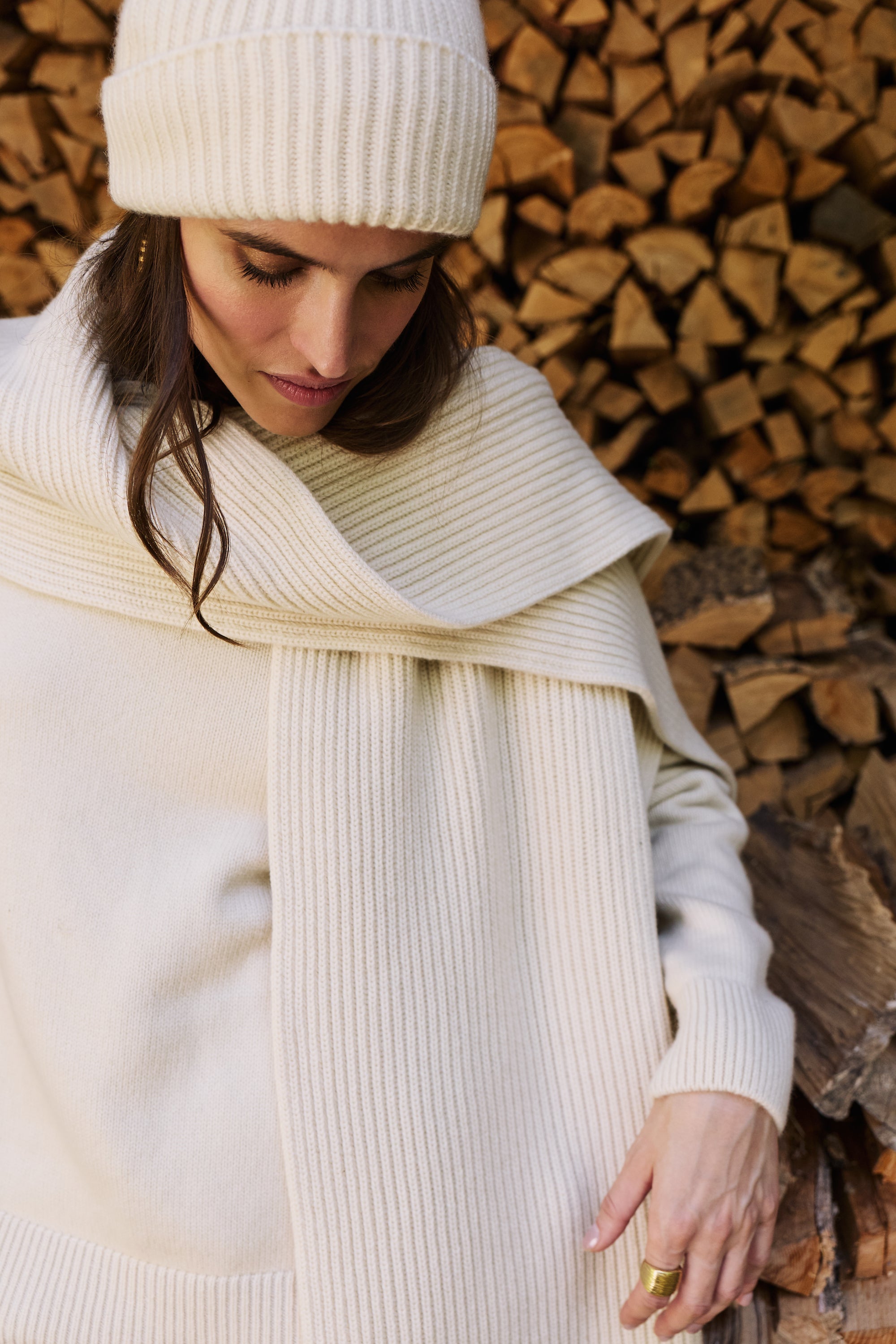 A woman wearing a cream ribbed knit beanie, oversized scarf, and matching sweater stands in front of a stacked woodpile.