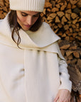 A woman wearing a cream ribbed knit beanie, oversized scarf, and matching sweater stands in front of a stacked woodpile.