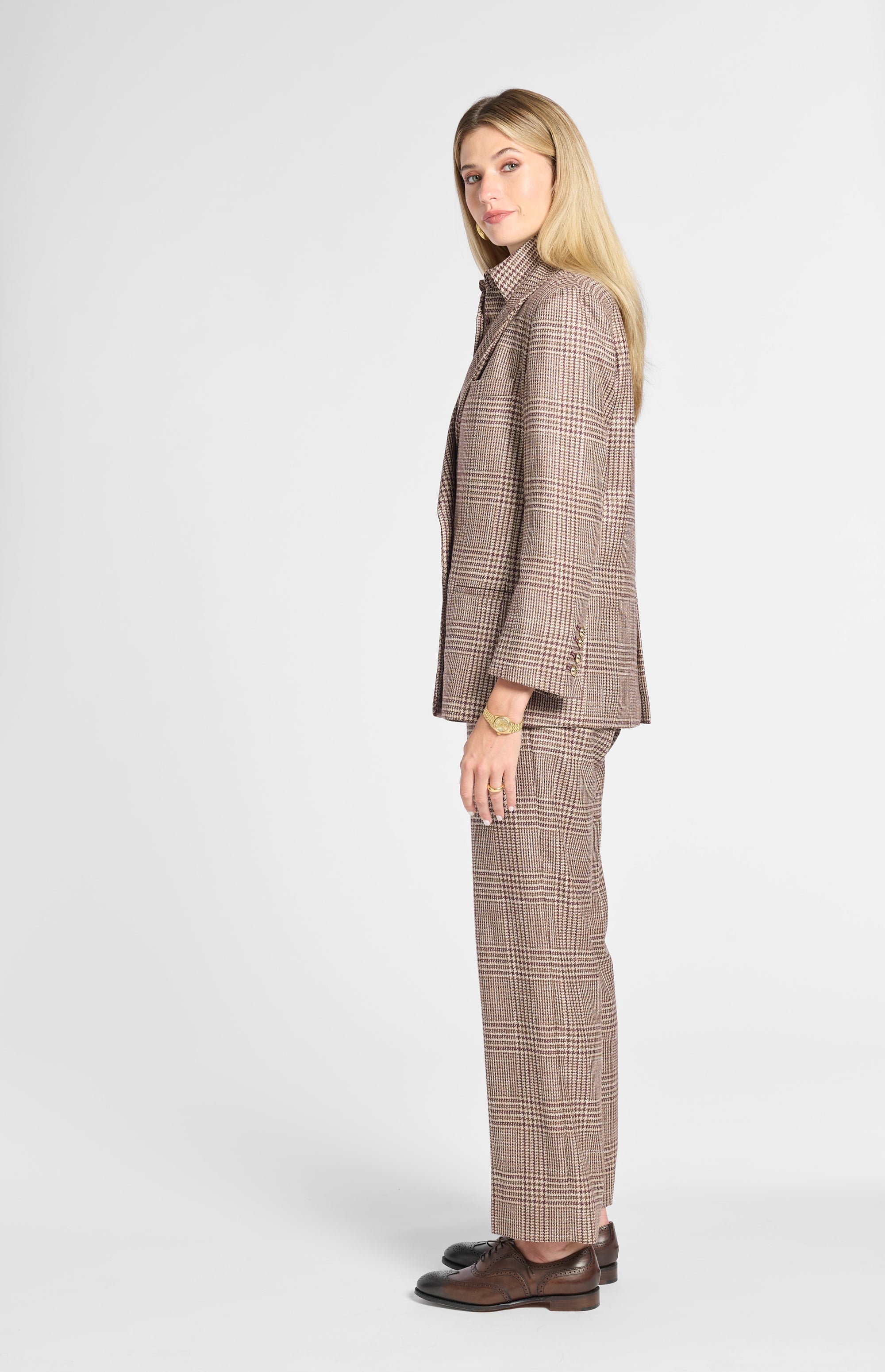 A woman modeling a brown and beige plaid suit, featuring a tailored blazer and matching trousers, paired with dark brown leather shoes against a light gray studio background.