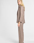 A woman modeling a brown and beige plaid suit, featuring a tailored blazer and matching trousers, paired with dark brown leather shoes against a light gray studio background.