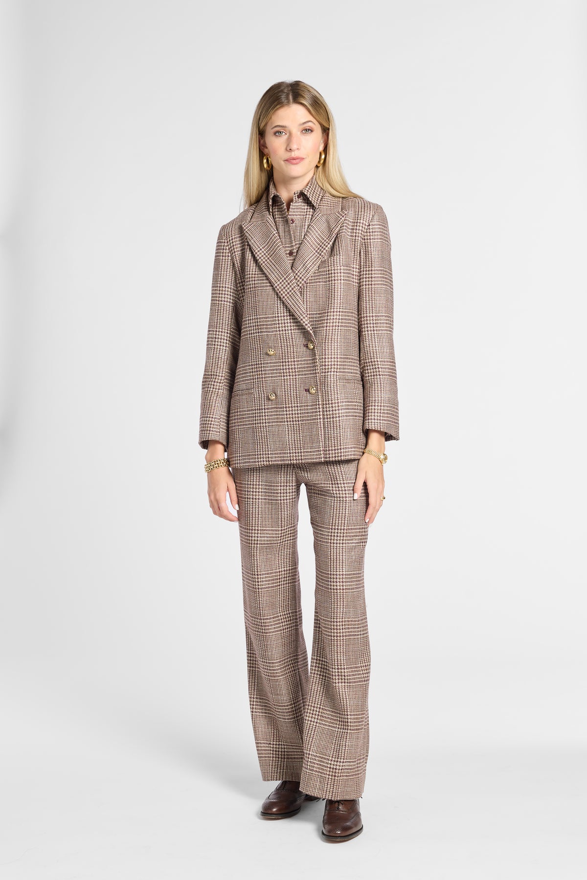 A woman modeling a matching brown and beige plaid double-breasted blazer and wide-leg trousers suit, paired with a collared shirt and brown leather shoes, against a light gray studio background.