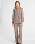 A woman modeling a matching brown and beige plaid double-breasted blazer and wide-leg trousers suit, paired with a collared shirt and brown leather shoes, against a light gray studio background.