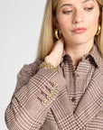 A close-up of a woman wearing a brown and beige houndstooth patterned blazer over a matching collared shirt, accessorized with gold hoop earrings and a gold chain bracelet, against a light gray background.