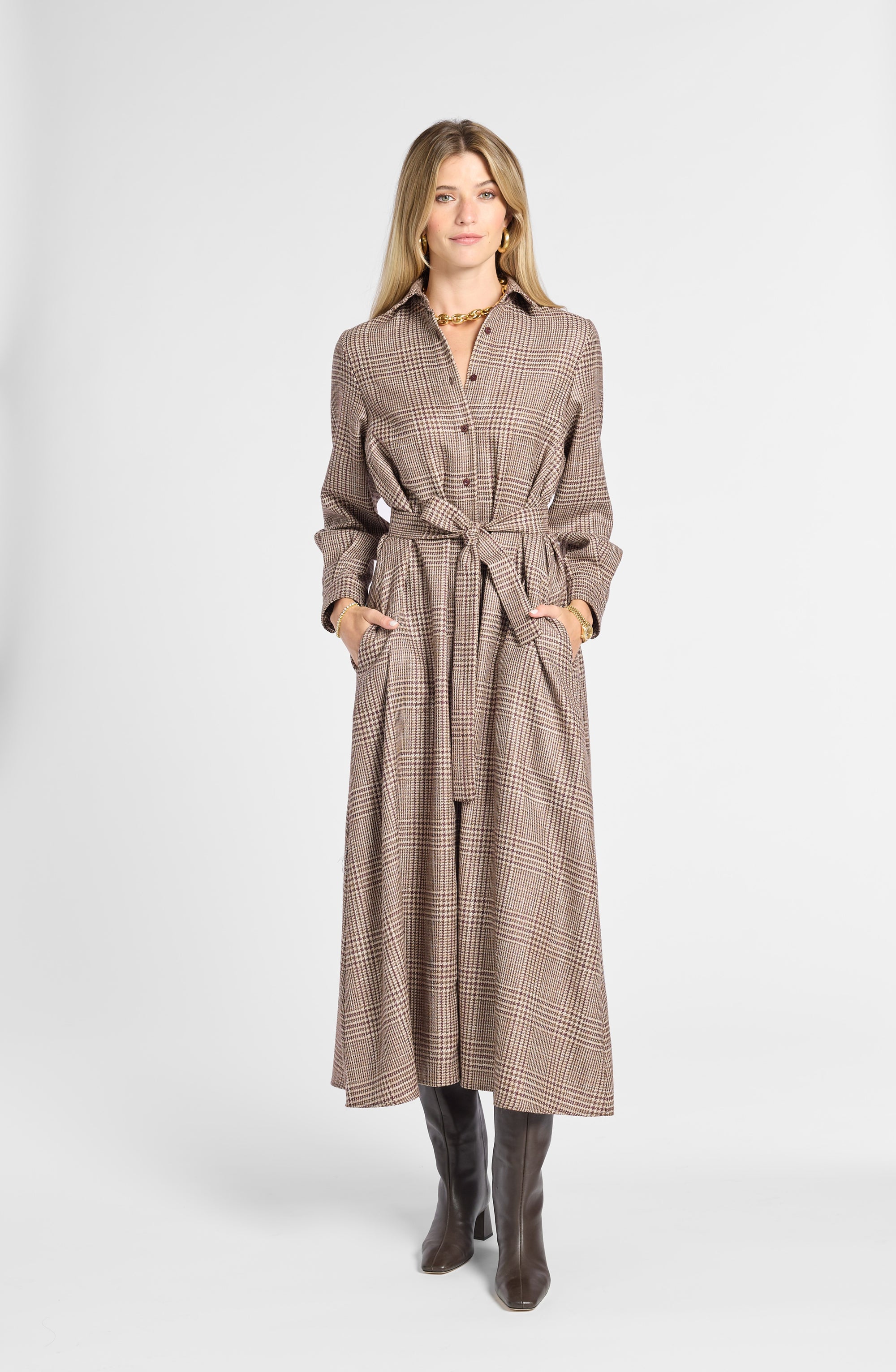 A woman modeling a long brown and beige houndstooth belted coat dress with pockets, paired with dark brown boots, against a light gray studio background.