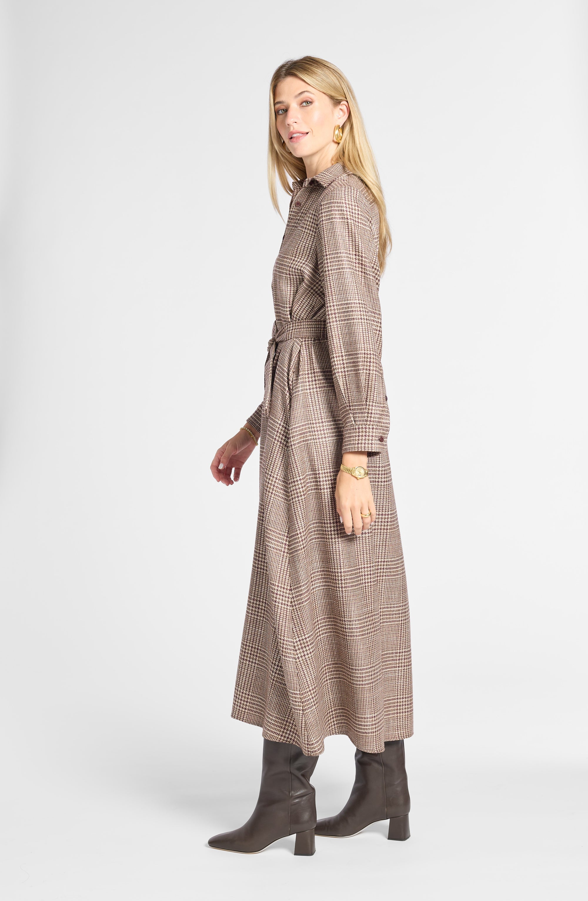 A woman modeling a long-sleeved, collared plaid midi dress in brown and beige tones, paired with dark brown heeled boots, against a light gray studio background.
