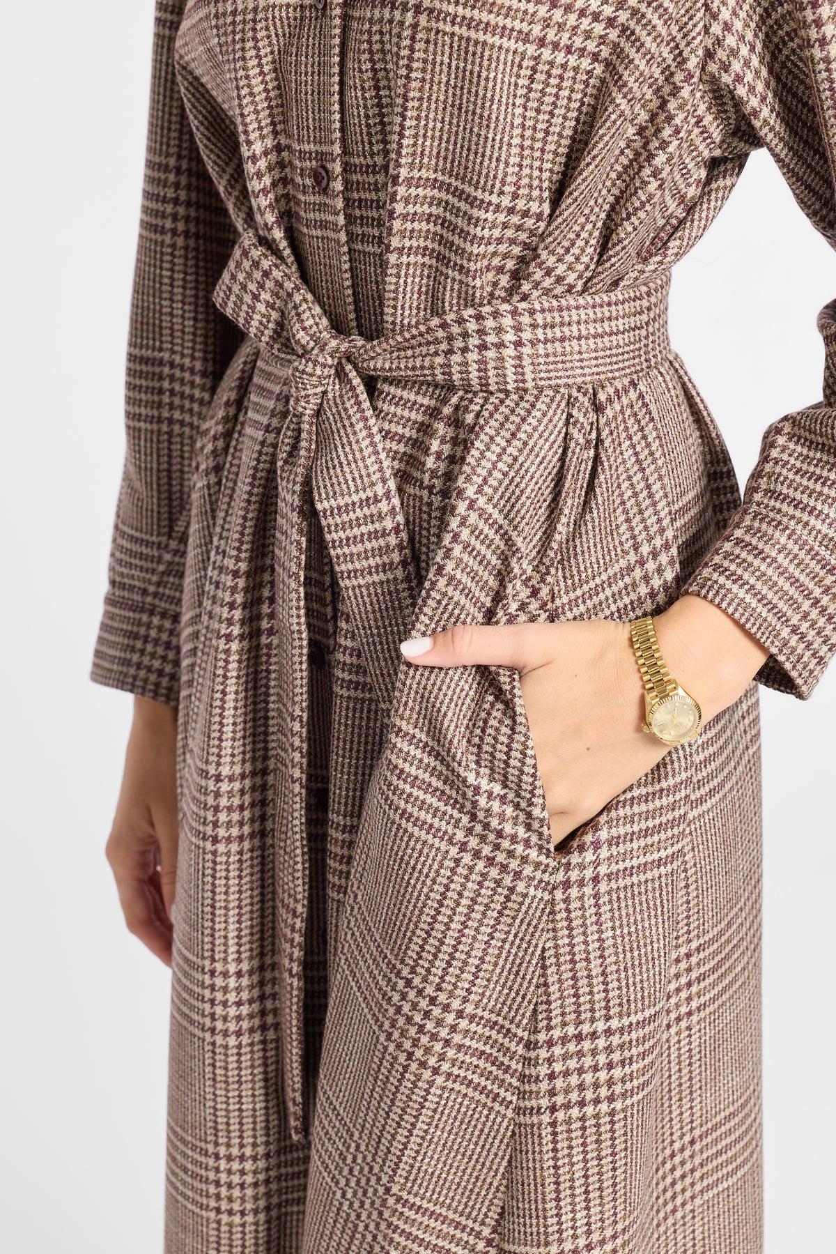 A close-up of a person wearing a brown and beige houndstooth patterned belted coat, with one hand in a pocket and a gold watch visible on the wrist, against a plain light background.
