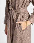 A close-up of a person wearing a brown and beige houndstooth patterned belted coat, with one hand in a pocket and a gold watch visible on the wrist, against a plain light background.