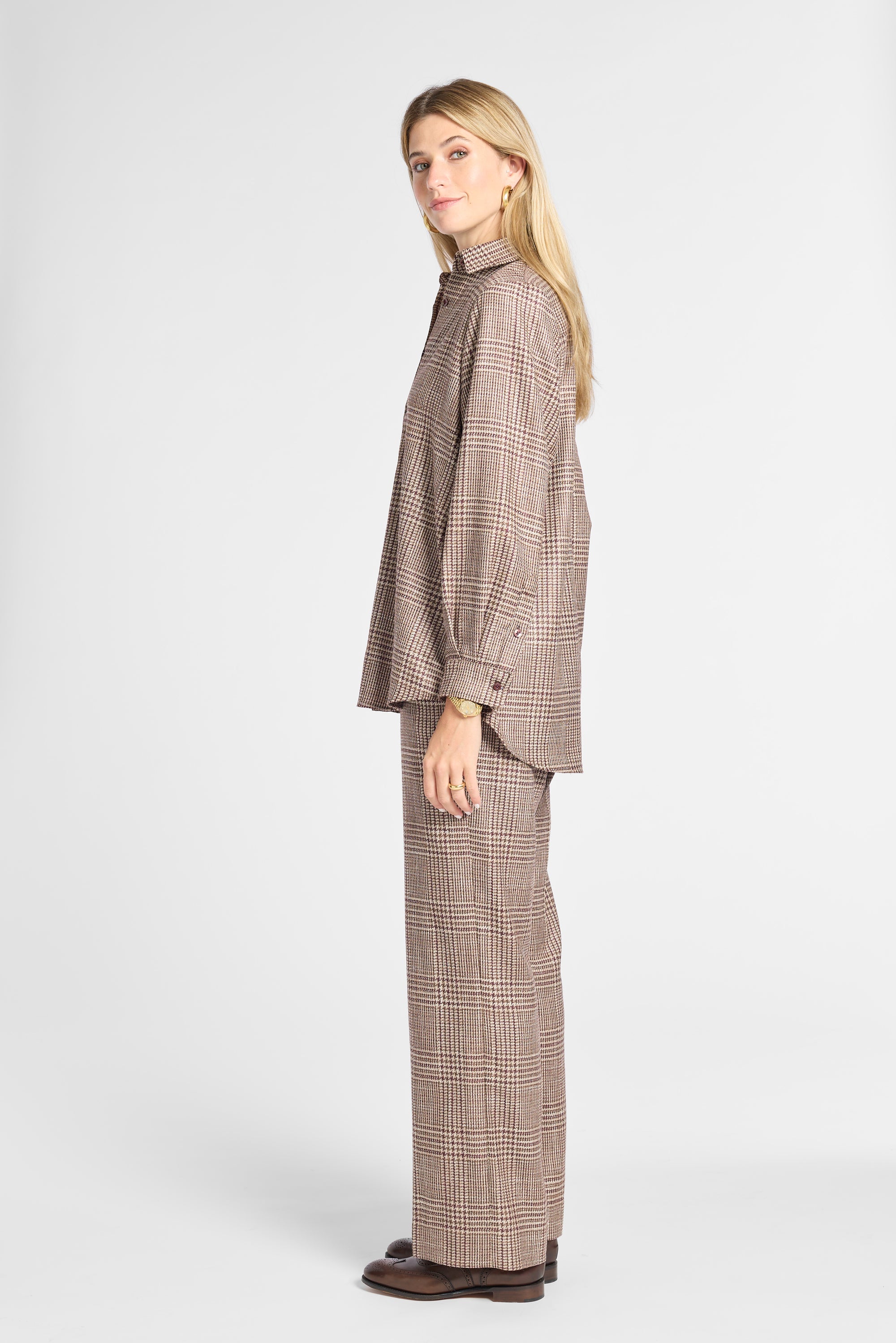 A woman modeling a matching brown and beige plaid two-piece suit, featuring a long-sleeve button-up shirt and wide-leg trousers, against a plain light gray studio background.