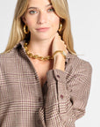 A close-up of a woman wearing a brown and beige houndstooth button-up shirt, styled with gold jewelry including a chunky gold chain necklace, gold hoop earrings, and a gold watch.