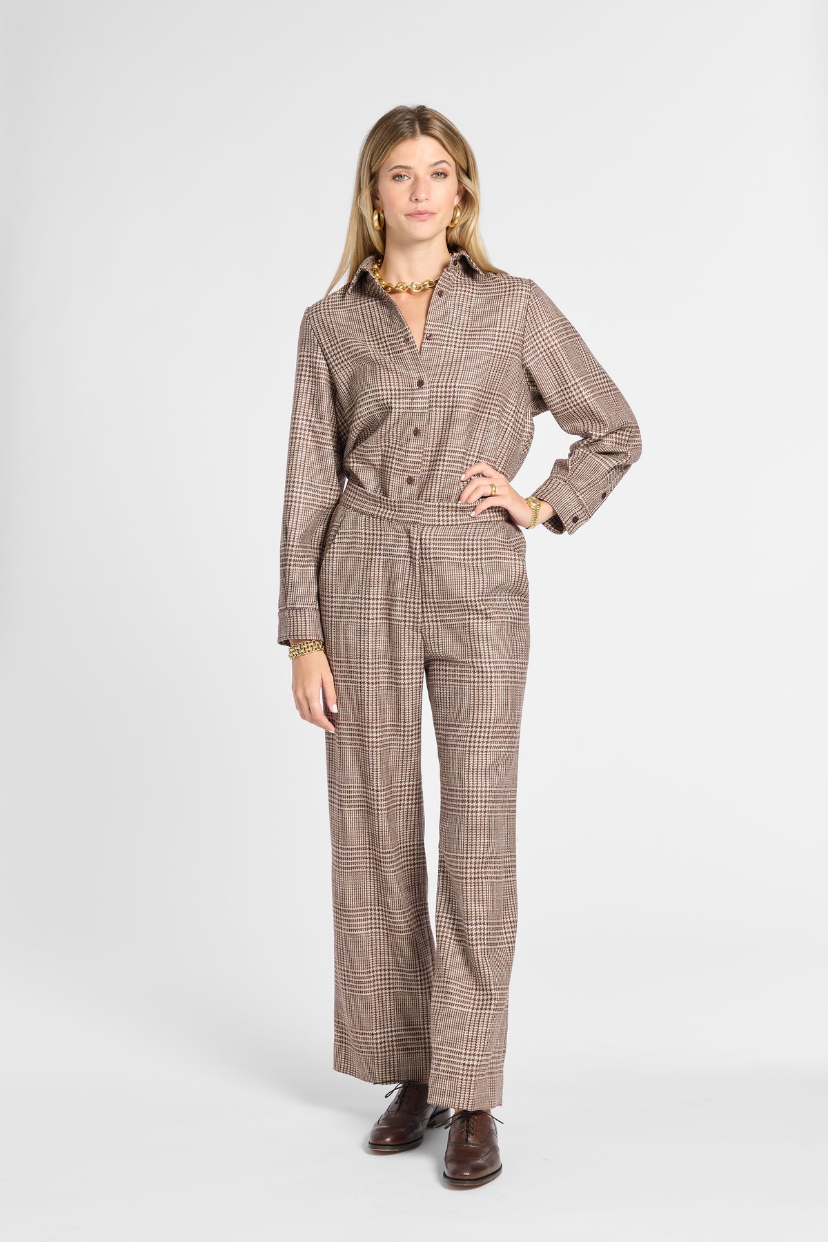 A woman modeling a brown and beige plaid long-sleeve button-up shirt and matching wide-leg trousers, styled with brown leather shoes and gold jewelry against a light gray studio background.