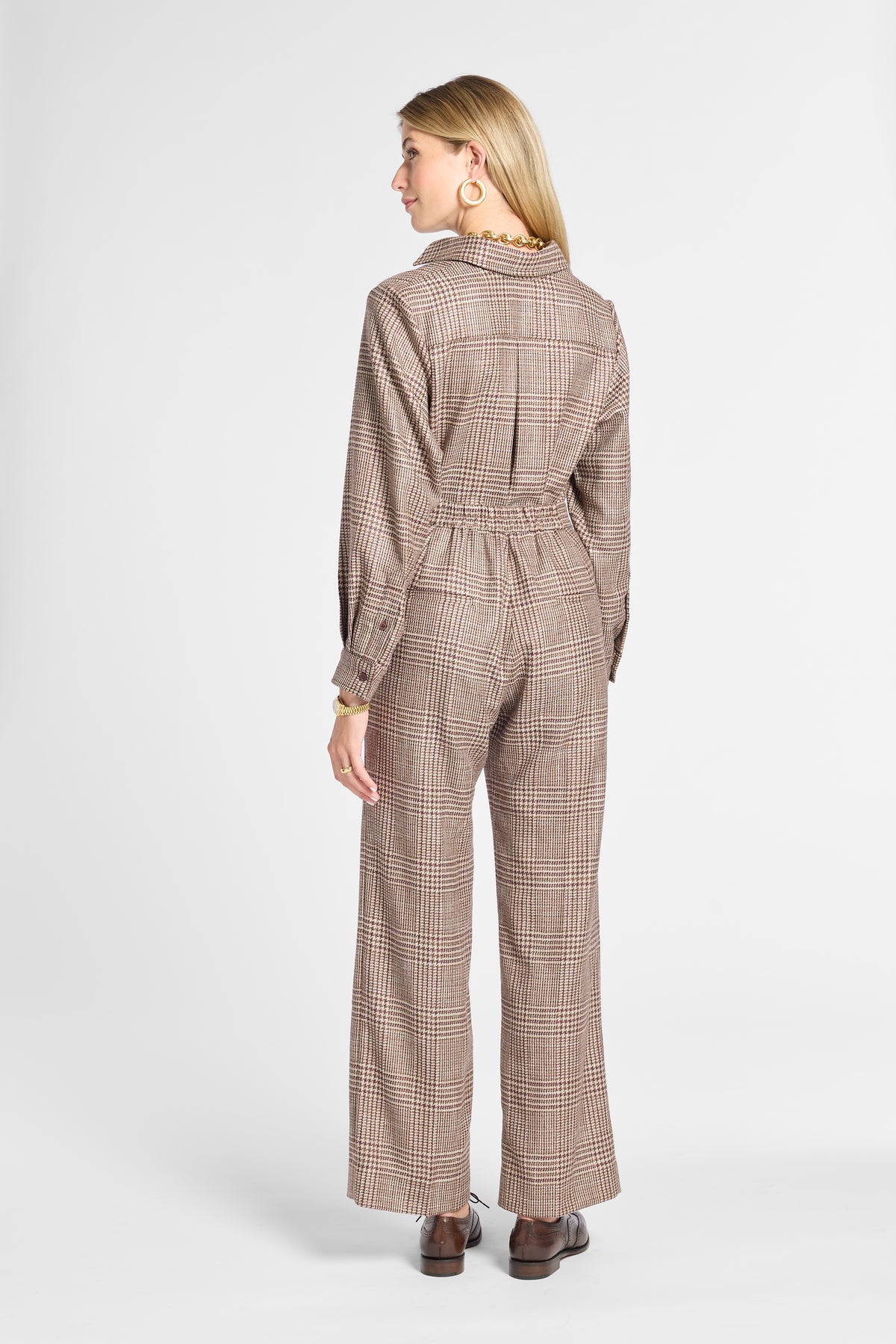 A woman shown from the back wearing a brown and beige plaid pair of pants and top with a high collar, long sleeves, and wide legs, paired with brown shoes, against a light gray studio background.