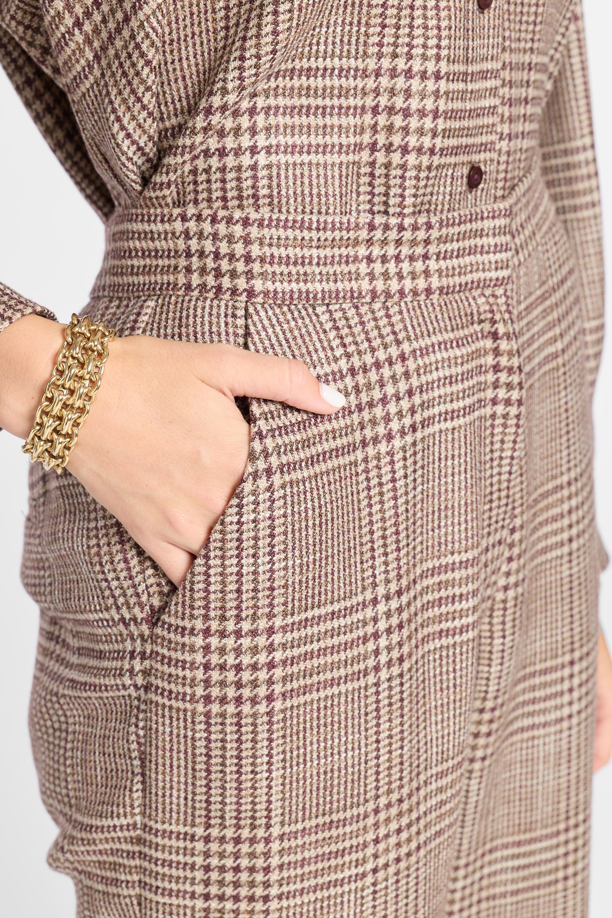 Close-up of a brown and beige plaid pair of pants with a hand tucked into the pocket, wearing a chunky gold chain bracelet.