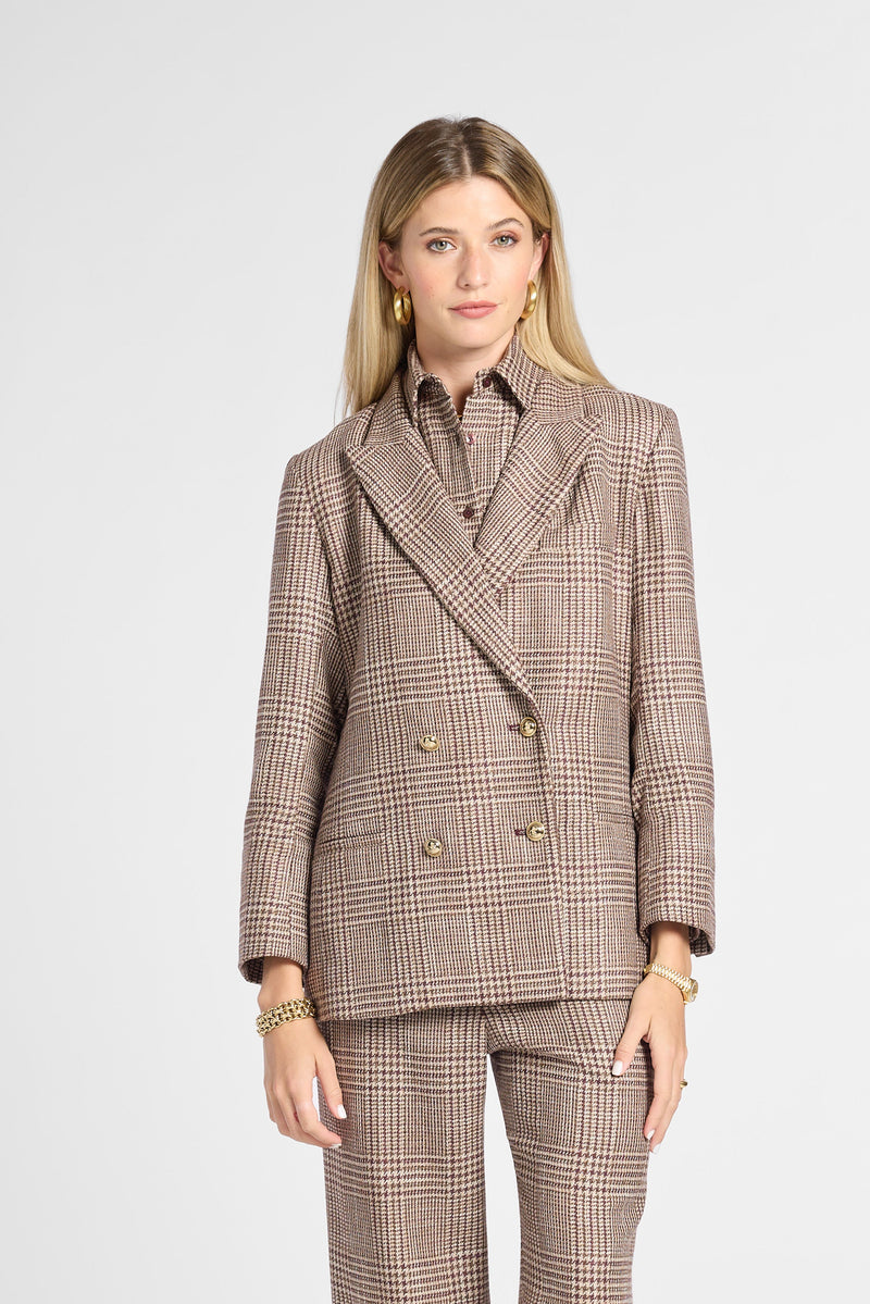 A woman modeling a brown and beige plaid double-breasted suit with matching trousers, worn with a collared shirt and brown leather shoes, against a light gray studio background.
