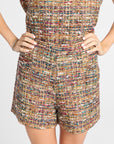 A close-up of a model wearing a strapless tweed romper with matching shorts. The fabric is a highly textured, multicolored bouclé weave containing shades of brown, beige, gold, and red.