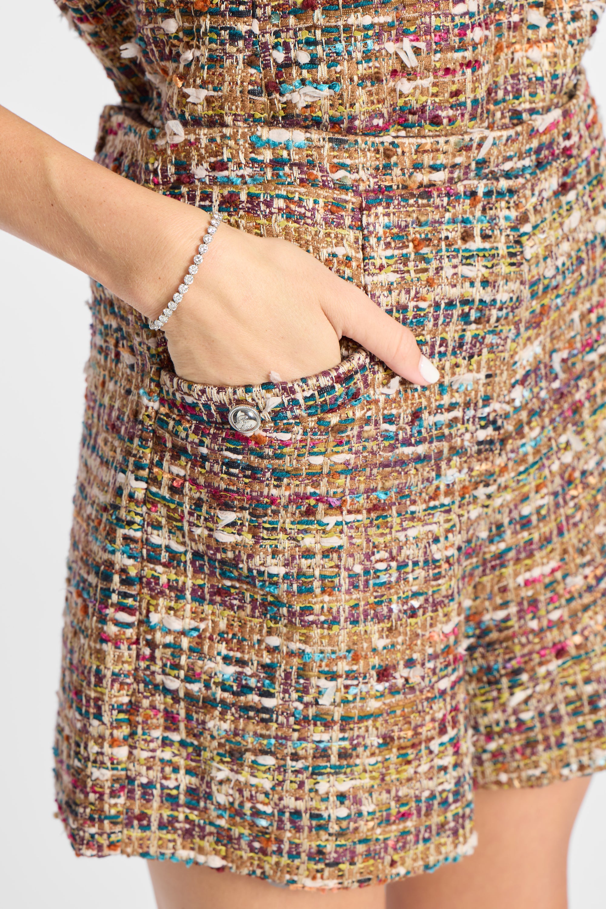 Close-up of a person wearing a multicolor tweed mini skirt with a side pocket and a silver beaded bracelet on the wrist, against a plain light background.