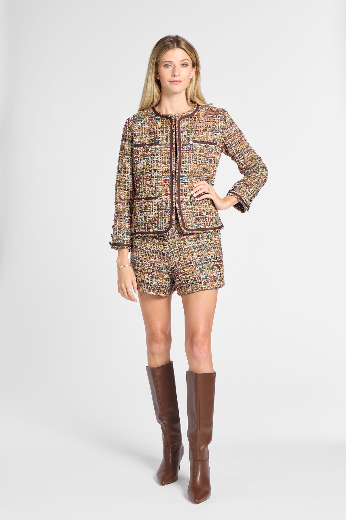 A woman models a matching tweed jacket and shorts set in a multicolor pattern with dark brown trim, paired with brown knee-high boots, against a light gray studio background.