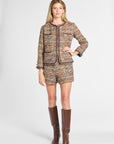 A woman models a matching tweed jacket and shorts set in a multicolor pattern with dark brown trim, paired with brown knee-high boots, against a light gray studio background.
