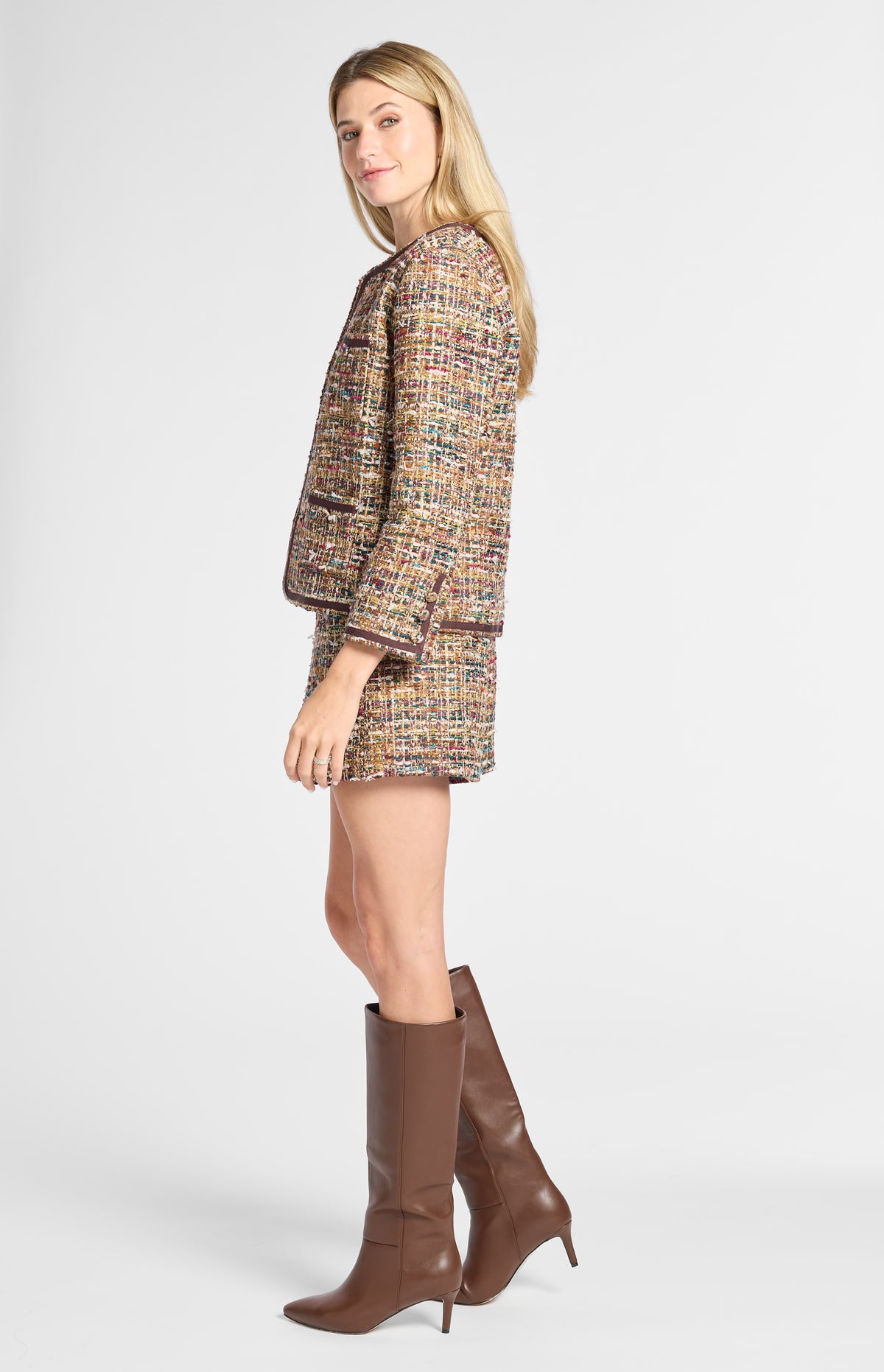 A woman modeling a tweed jacket and matching mini skirt in a multicolor woven pattern, paired with brown knee-high leather boots, against a light gray studio background.
