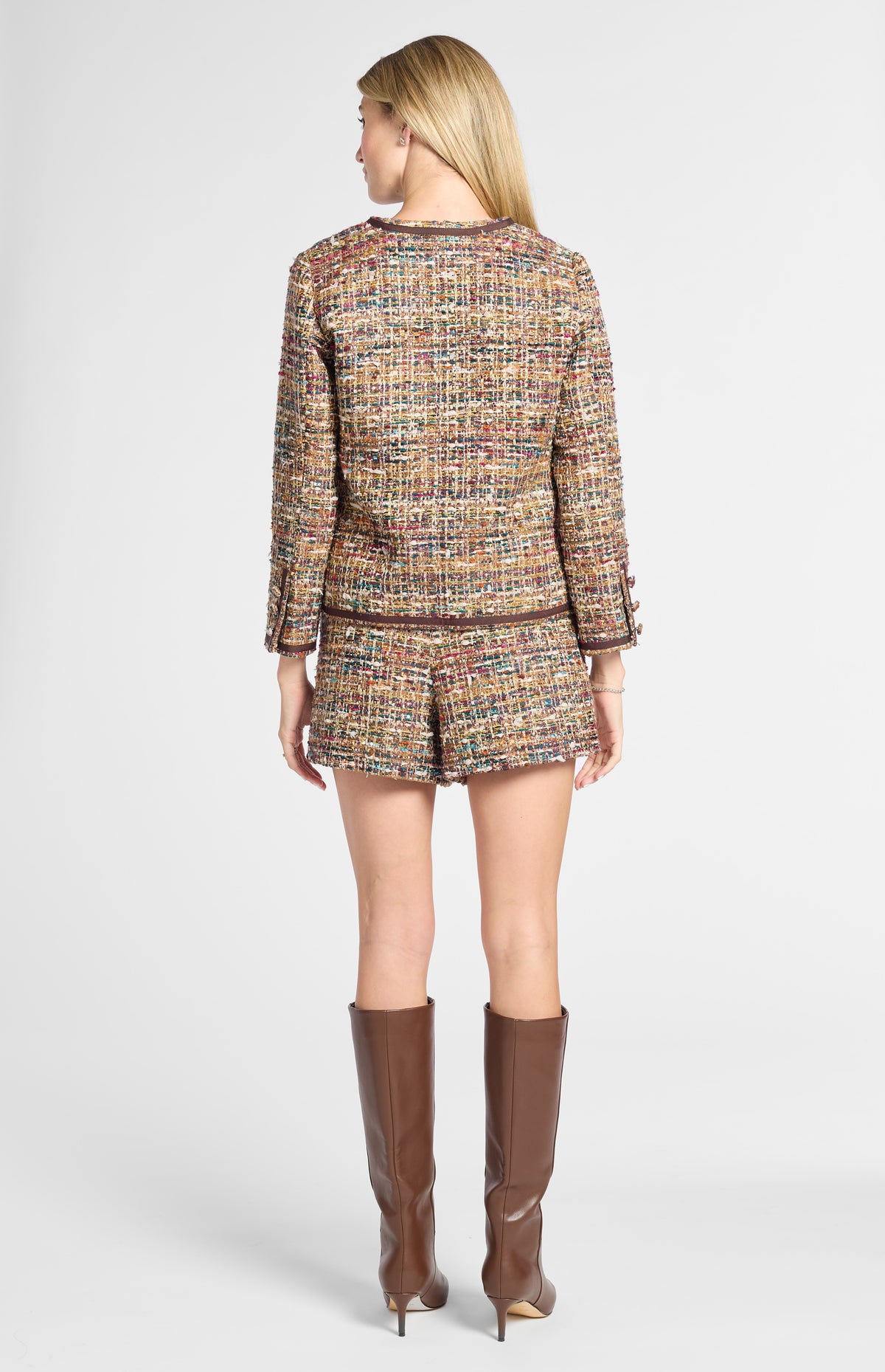 A woman viewed from the back wearing a tweed jacket and matching shorts in a multicolor woven pattern, paired with tall brown knee-high boots, against a light gray studio background.