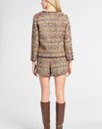 A woman viewed from the back wearing a tweed jacket and matching shorts in a multicolor woven pattern, paired with tall brown knee-high boots, against a light gray studio background.
