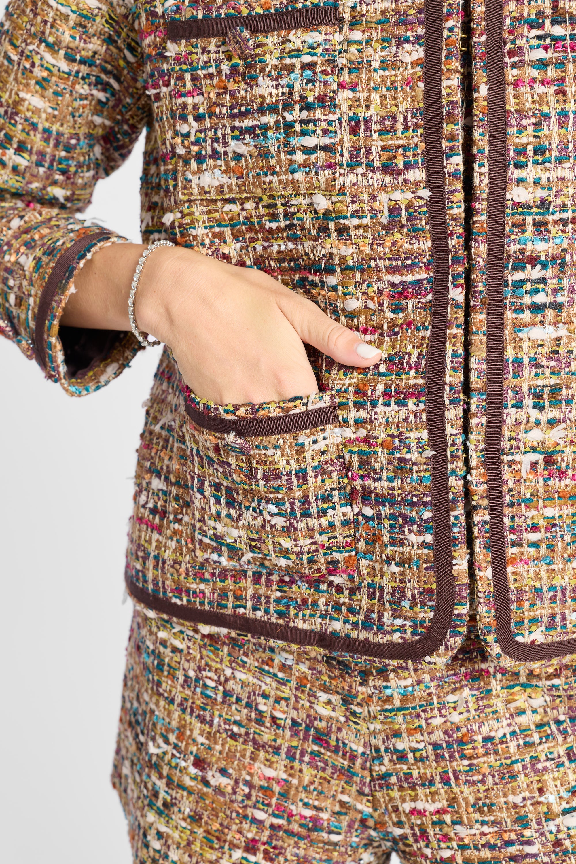 Close-up of a tweed blazer and matching trousers in a multicolor bouclé weave with dark brown trim, shown on a model with a hand in the pocket.