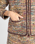 Close-up of a tweed blazer and matching trousers in a multicolor bouclé weave with dark brown trim, shown on a model with a hand in the pocket.