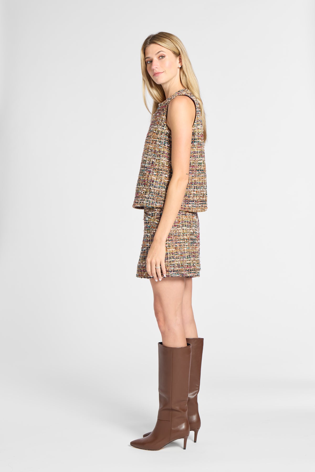 A woman modeling a sleeveless tweed mini dress with a matching mini skirt in a multicolor bouclé pattern, paired with brown knee-high leather boots, against a light gray studio background.