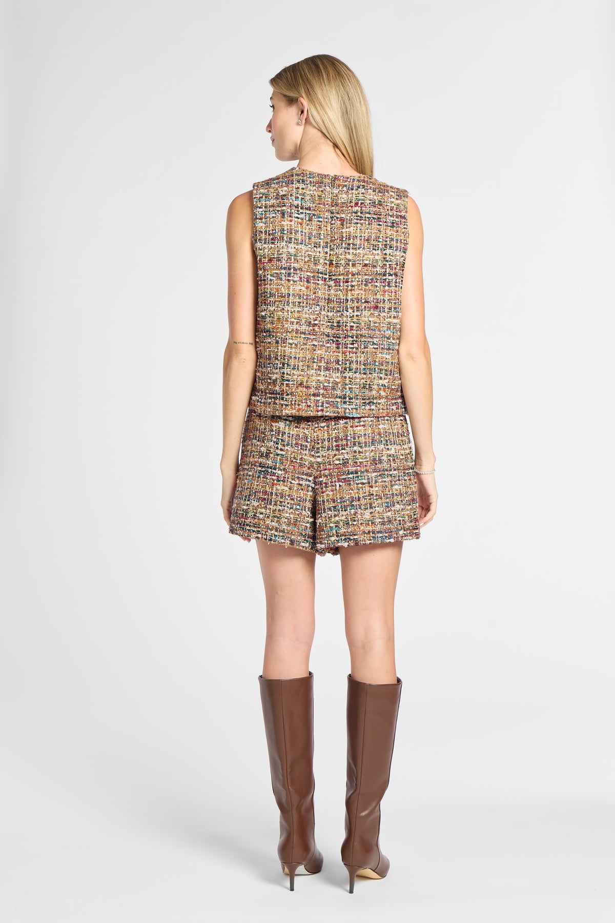 A woman shown from the back wearing a sleeveless tweed vest and matching shorts in a multicolor woven pattern, paired with brown knee-high boots, against a light gray studio background.