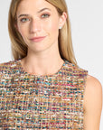 A close-up of a woman wearing a sleeveless textured tweed top with a multicolor pattern of beige, blue, pink, and gold threads, set against a plain light gray background.