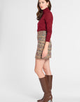 A woman modeling a burgundy long-sleeve turtleneck sweater, a multicolor tweed mini skirt, and brown knee-high leather boots, shown from a side profile against a light gray studio background.