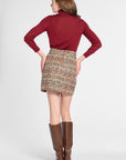 A woman shown from the back wearing a burgundy long-sleeve turtleneck top, a multicolor tweed mini skirt, and brown knee-high boots, against a light gray studio background.
