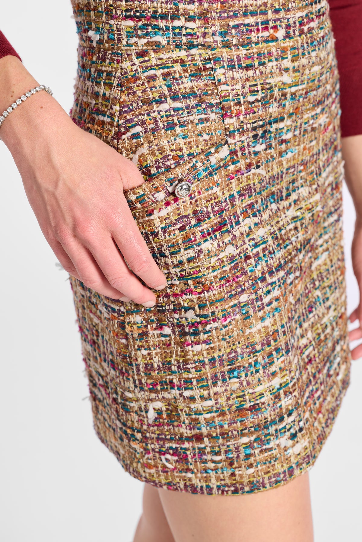 Close-up of a person wearing a textured tweed mini skirt with a multicolor pattern of gold, beige, red, and blue threads, featuring a silver button closure.