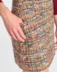 Close-up of a person wearing a textured tweed mini skirt with a multicolor pattern of gold, beige, red, and blue threads, featuring a silver button closure.