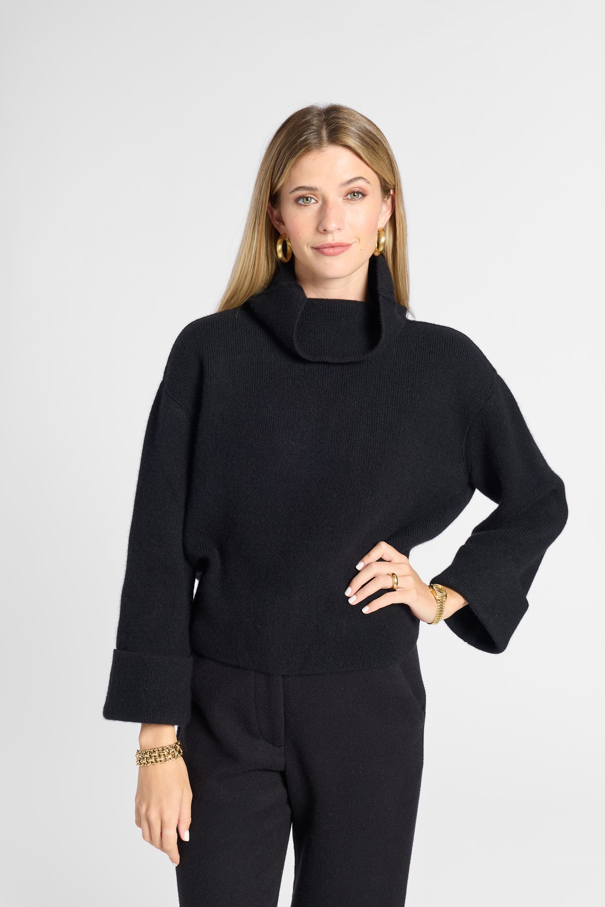A woman modeling a black knit turtleneck sweater with wide three-quarter sleeves and black trousers, posed against a light gray studio background.