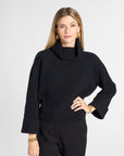 A woman modeling a black knit turtleneck sweater with wide three-quarter sleeves and black trousers, posed against a light gray studio background.