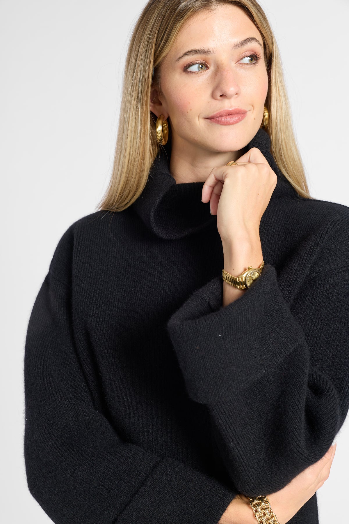 A woman wearing a black ribbed knit mock neck sweater, styled with gold hoop earrings, a gold watch, and a gold bracelet, against a light gray studio background.