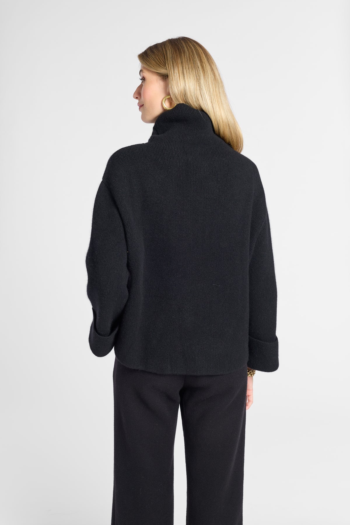 A woman shown from the back wearing a black knit turtleneck sweater and black wide-leg trousers against a light gray studio background.