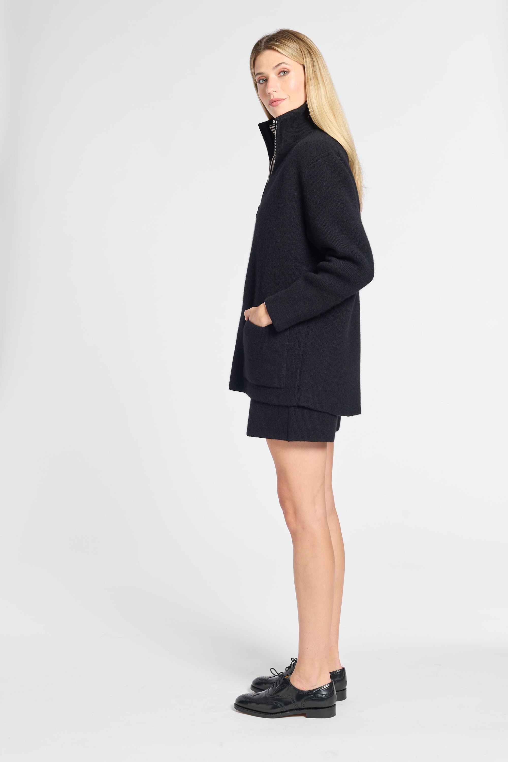 A blonde model wearing a black oversized zip-up coat, matching black shorts, and black leather loafers, standing in profile against a light gray studio background.