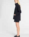 A blonde model wearing a black oversized zip-up coat, matching black shorts, and black leather loafers, standing in profile against a light gray studio background.