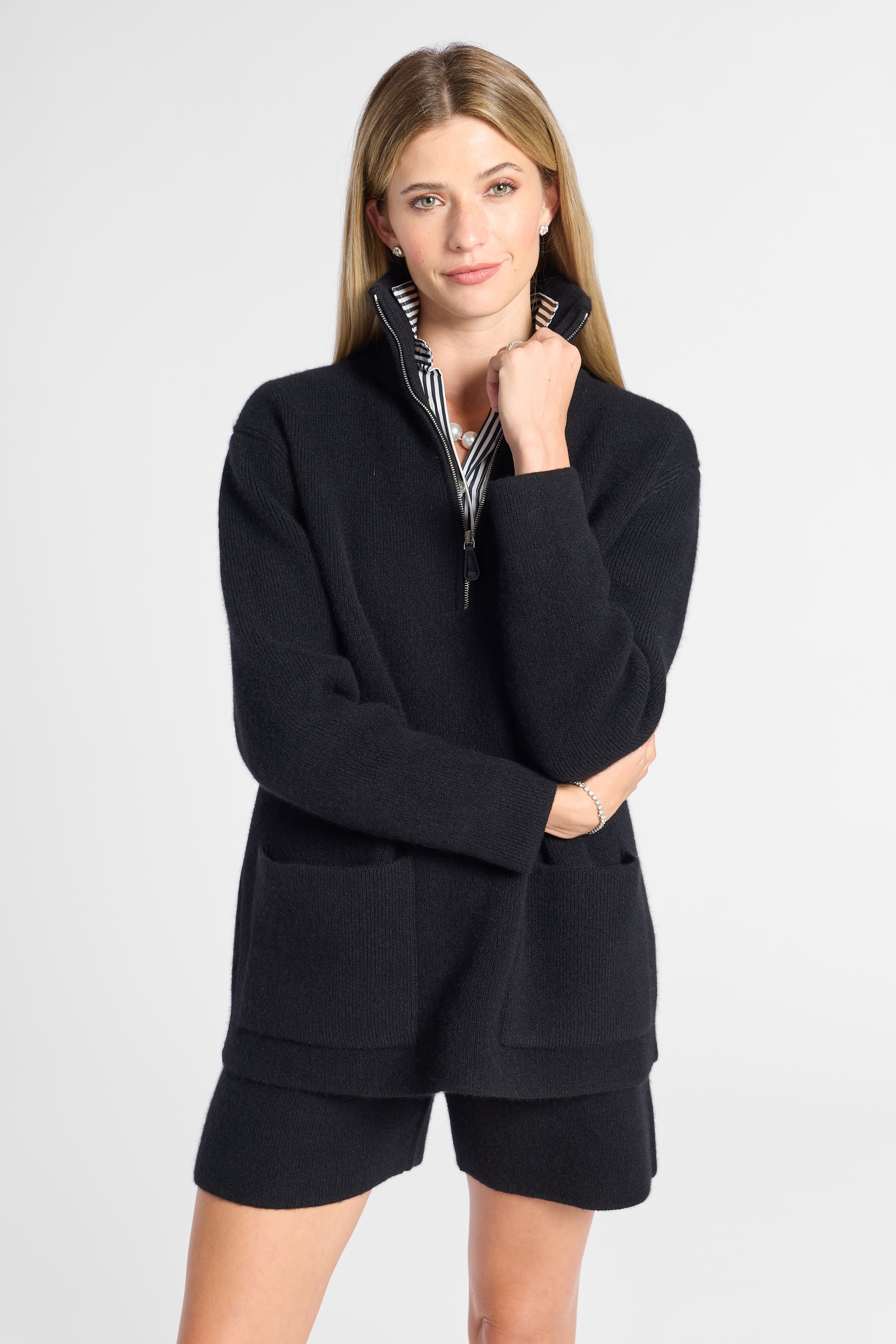 A woman modeling a black knit quarter-zip sweater with a striped inner collar and matching black shorts against a light gray studio background.