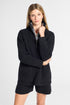 A woman modeling a black knit quarter-zip sweater with a striped inner collar and matching black shorts against a light gray studio background.