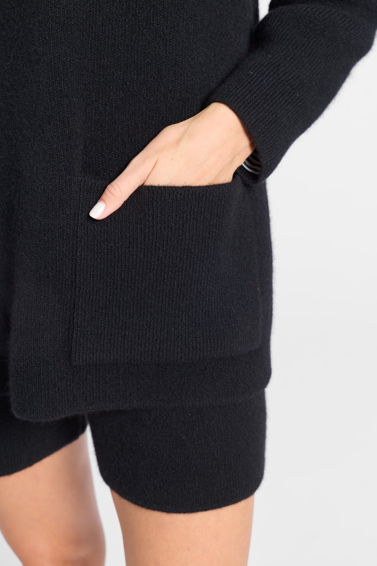Close-up of a person wearing a black ribbed knit sweater with a large front patch pocket and matching black knit shorts, with one hand tucked into the pocket against a light gray background.